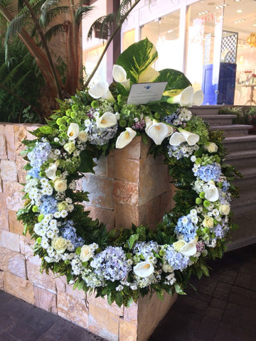 Corona fúnebre hecha con flores naturales blancas y celestes con follajes verdes.
