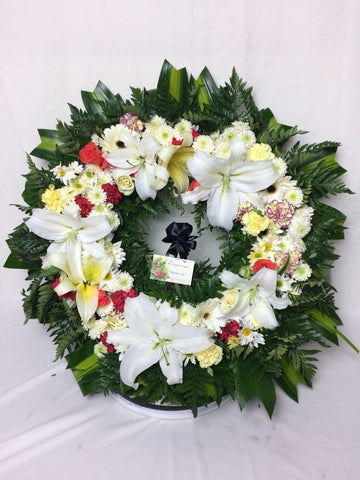 Corona para difunto hecha con lirios, rosas y variedad de follajes verdes.