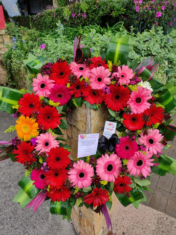 Elegante corona fúnebre con gerberas rosadas y rojas con follajes verdes.