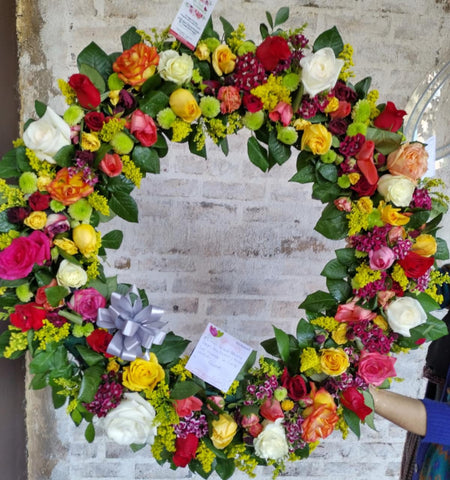 Corona de flores naturales con 4 doces de rosas de varios colores y follajes verdes.