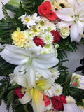 Corona fúnebre hecha con lirios, rosas y variedad de follajes verdes.