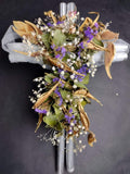 Cruz plateada con flores artificiales color lilas y verdes.