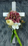 Elegante cruz de madera con flores artificiales blancas, moradas y hojas verdes.